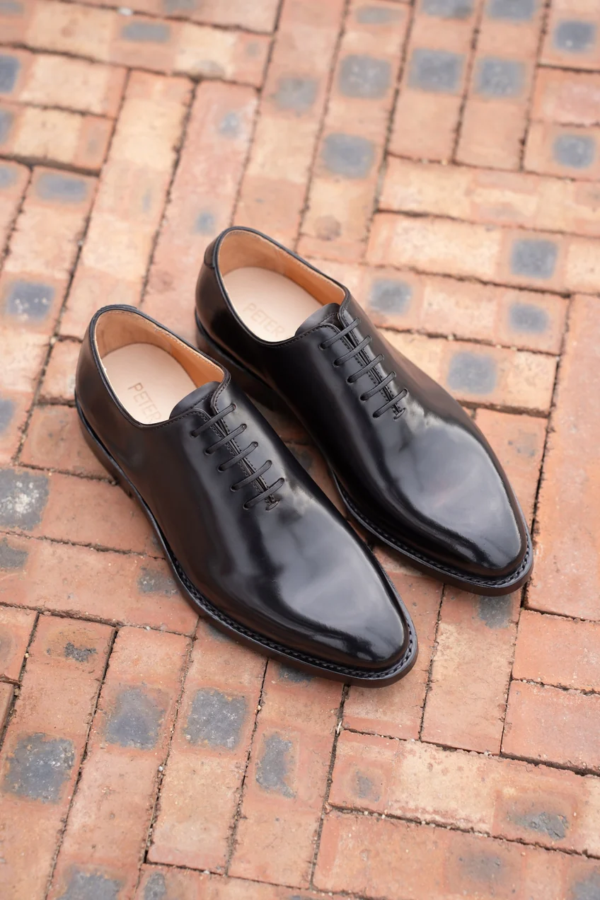 A pair of polished black cordovan shoes with a pointed toe and six eyelets for laces.