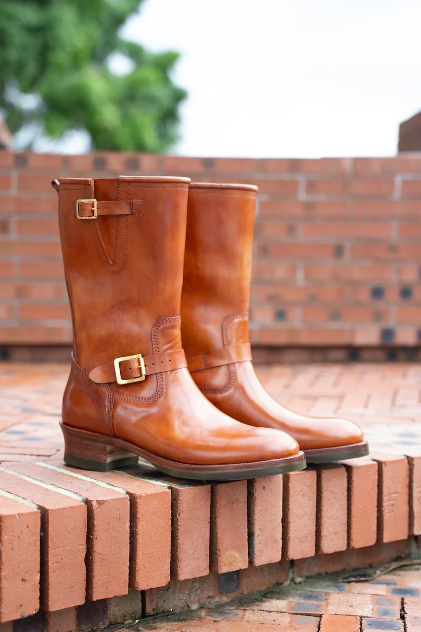A pair of polished brown cordovan leather boots with gold buckles and a stacked heel.