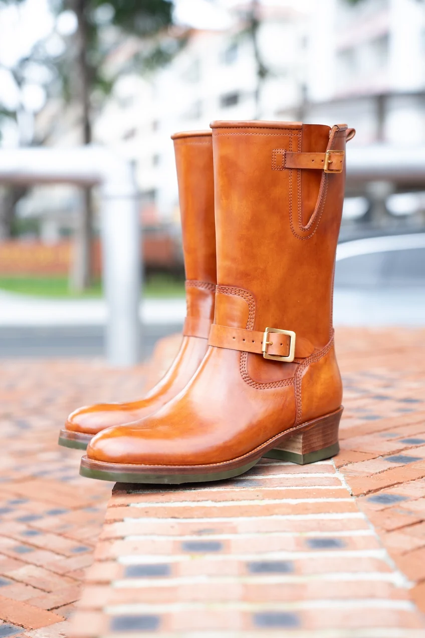 A pair of polished tan leather boots with a buckle detail and a stacked heel.