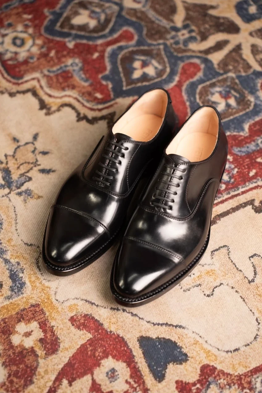 A pair of polished black cordovan shoes with a cap toe design and lace-up closure.
