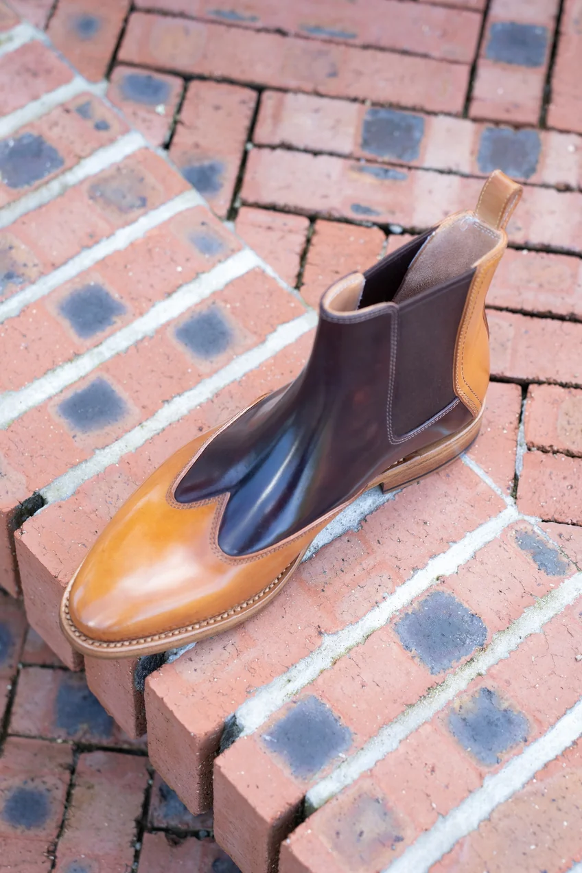 A brown and tan leather Chelsea boot with a pointed toe rests on a brick surface.