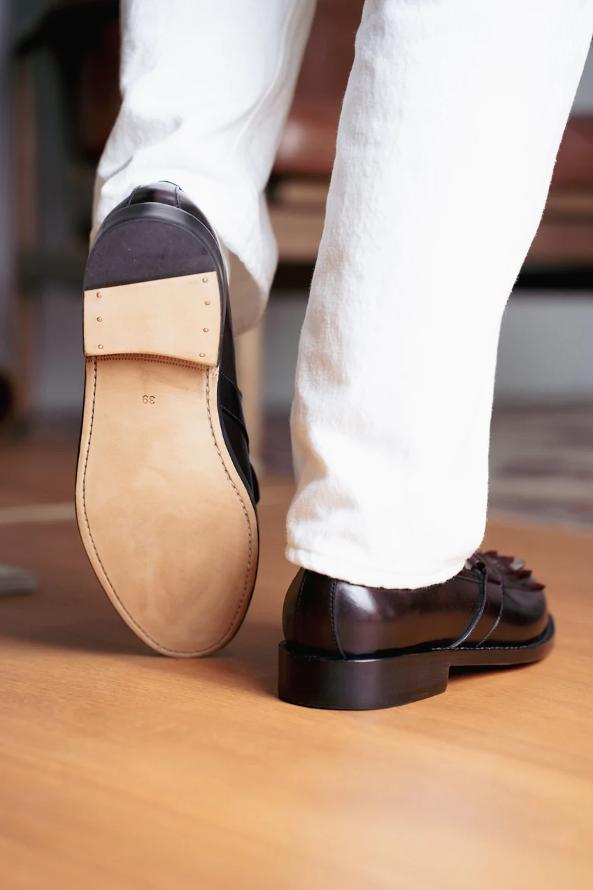 A close-up of a brown leather shoe with a smooth sole and visible stitching, worn with white pants.
