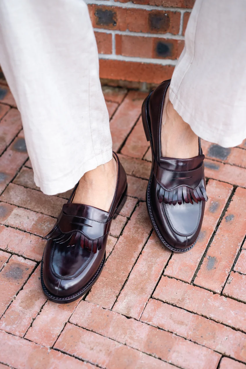 A pair of dark burgundy cordovan shoes with a fringed detail on the front, worn with light-colored pants.