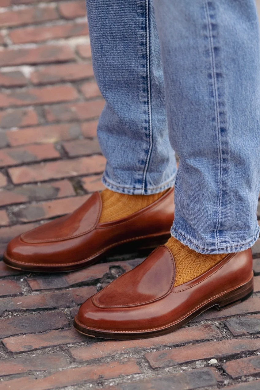 Brown cordovan loafers paired with mustard-colored socks and blue jeans.