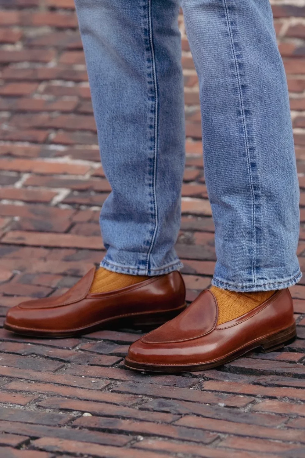 A close-up of brown leather slip-on shoes worn with blue jeans and mustard-colored socks.