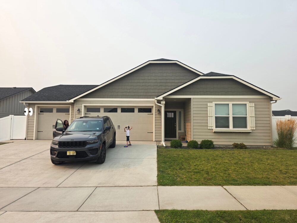 A black SUV is parked in front of a light gray house with white shutters, while a woman stands by the open driver's door and a child plays nearby.