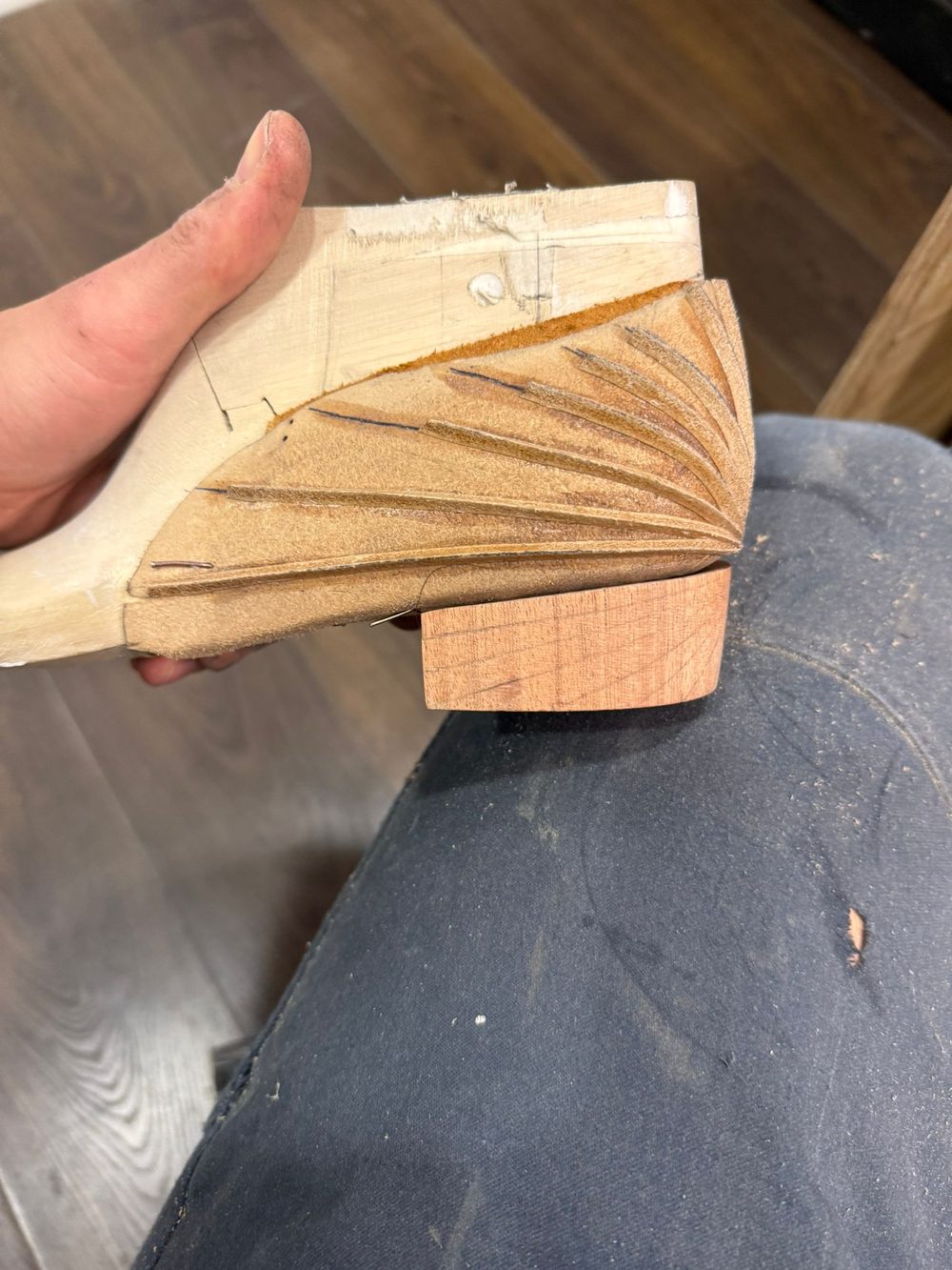 A close-up of a hand holding a carved wooden shoe heel with a textured design and a small block of wood attached.