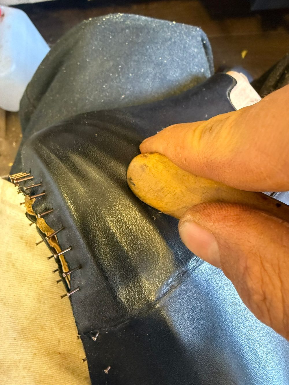 A hand holds a wooden tool against a black leather shoe upper, with visible metal pins along the edge.
