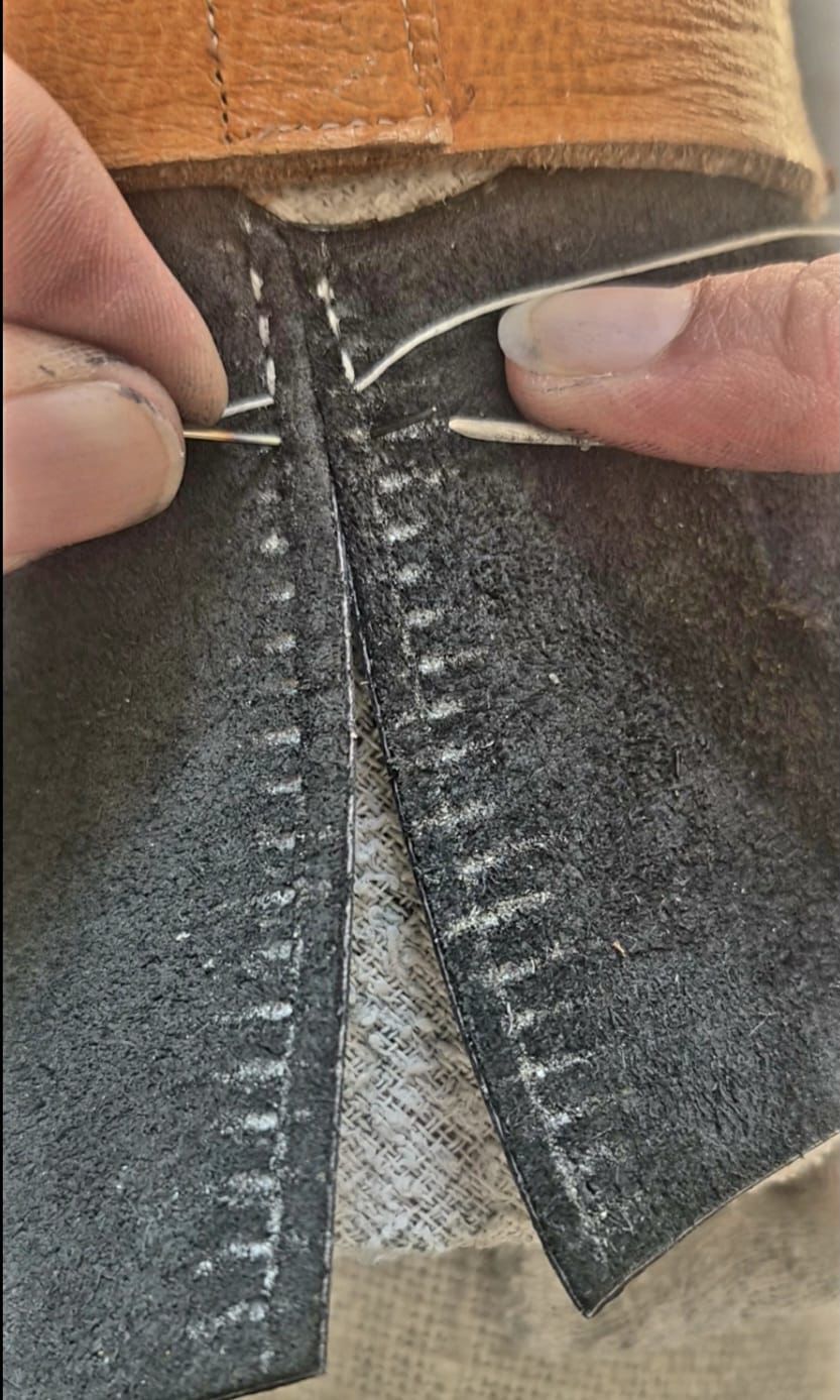 A close-up of hands stitching together two pieces of dark leather with a needle and thread.