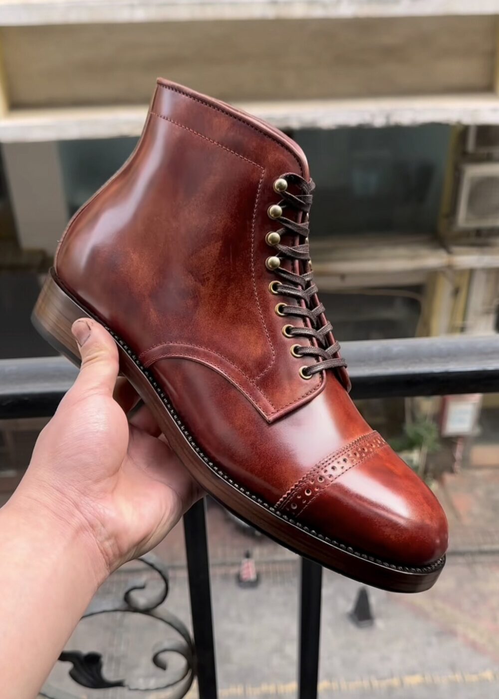 A hand holds a polished, brown cordovan leather boot with decorative stitching and metal eyelets.