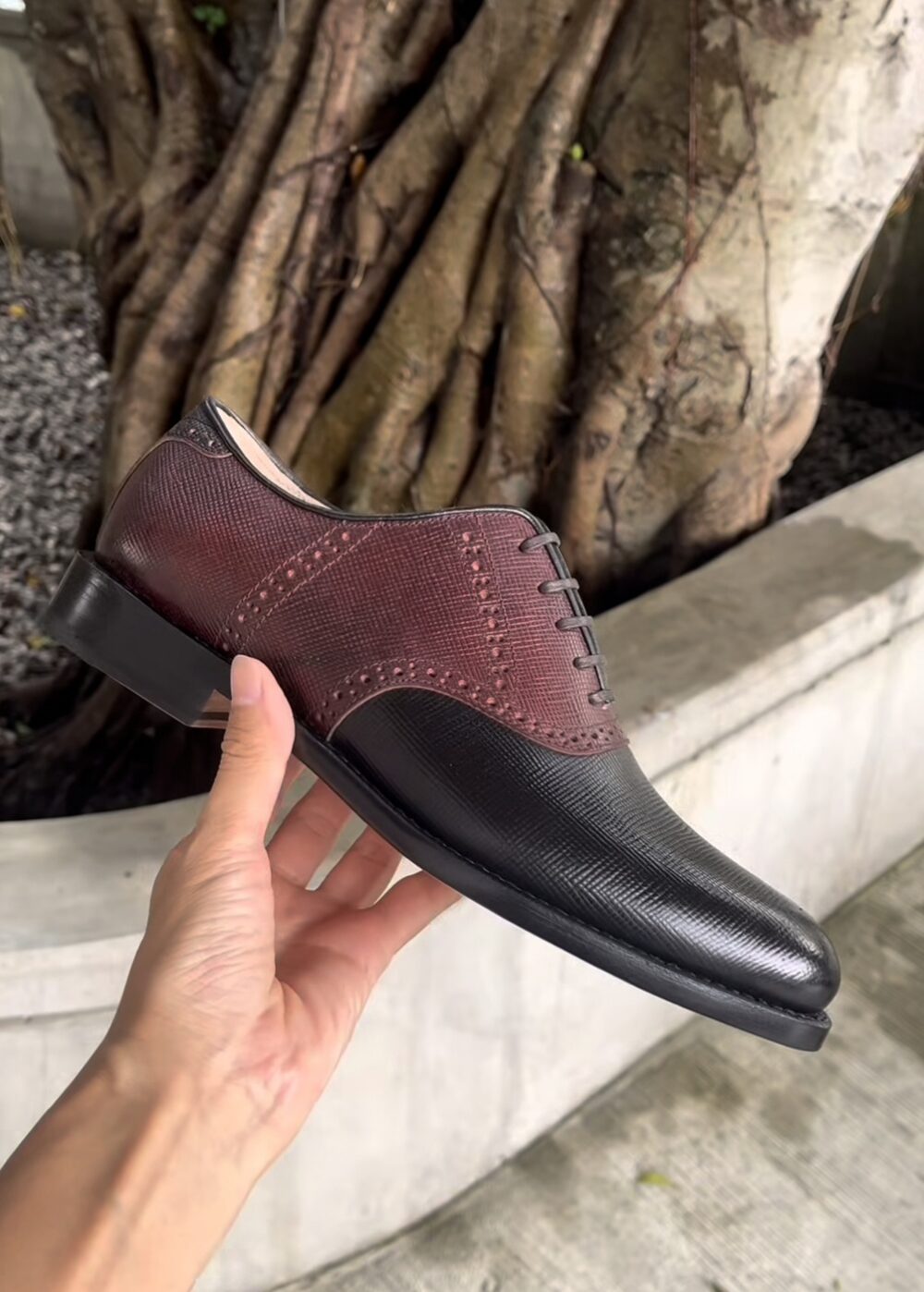 A hand holds a stylish shoe featuring a burgundy textured upper and a black smooth leather toe.