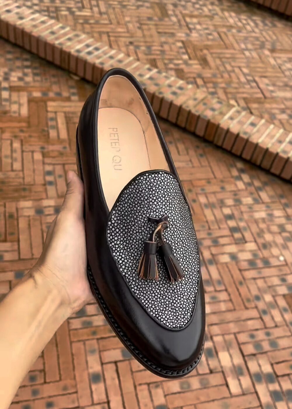 A black leather shoe with a textured grey fabric panel and tassel detail is being held by a hand.