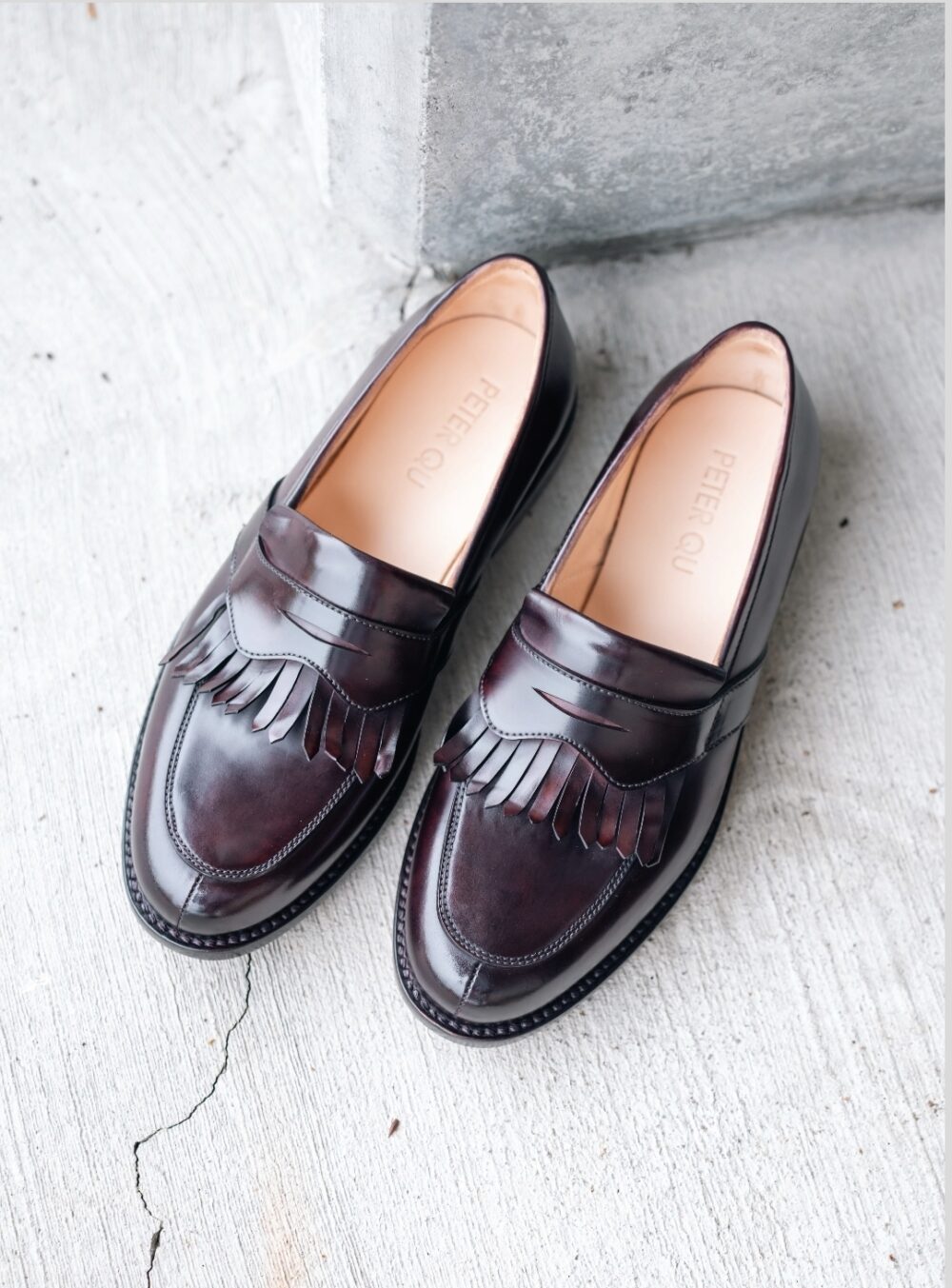 A pair of dark burgundy cordovan shoes with a fringed design on the front, labeled "PETER QU."