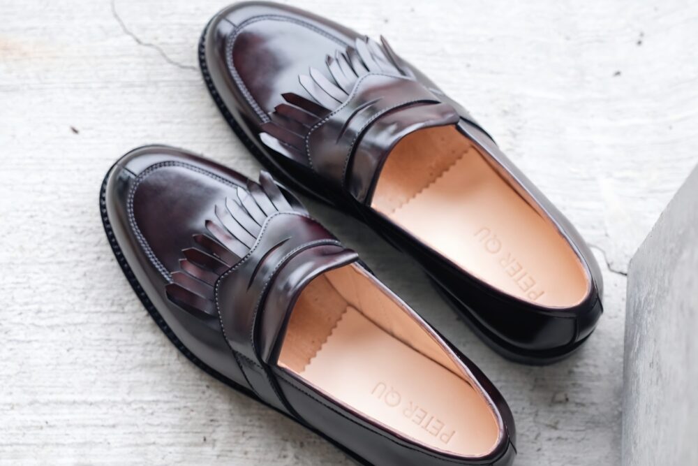 A pair of dark burgundy leather loafers with fringed detailing and a smooth tan interior, labeled "PETER QU."