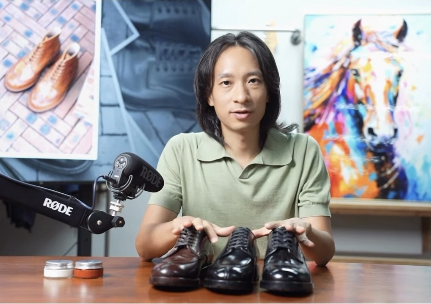 A person with long hair sits at a table, holding a brown shoe in one hand and a black shoe in the other, wearing a light green shirt.