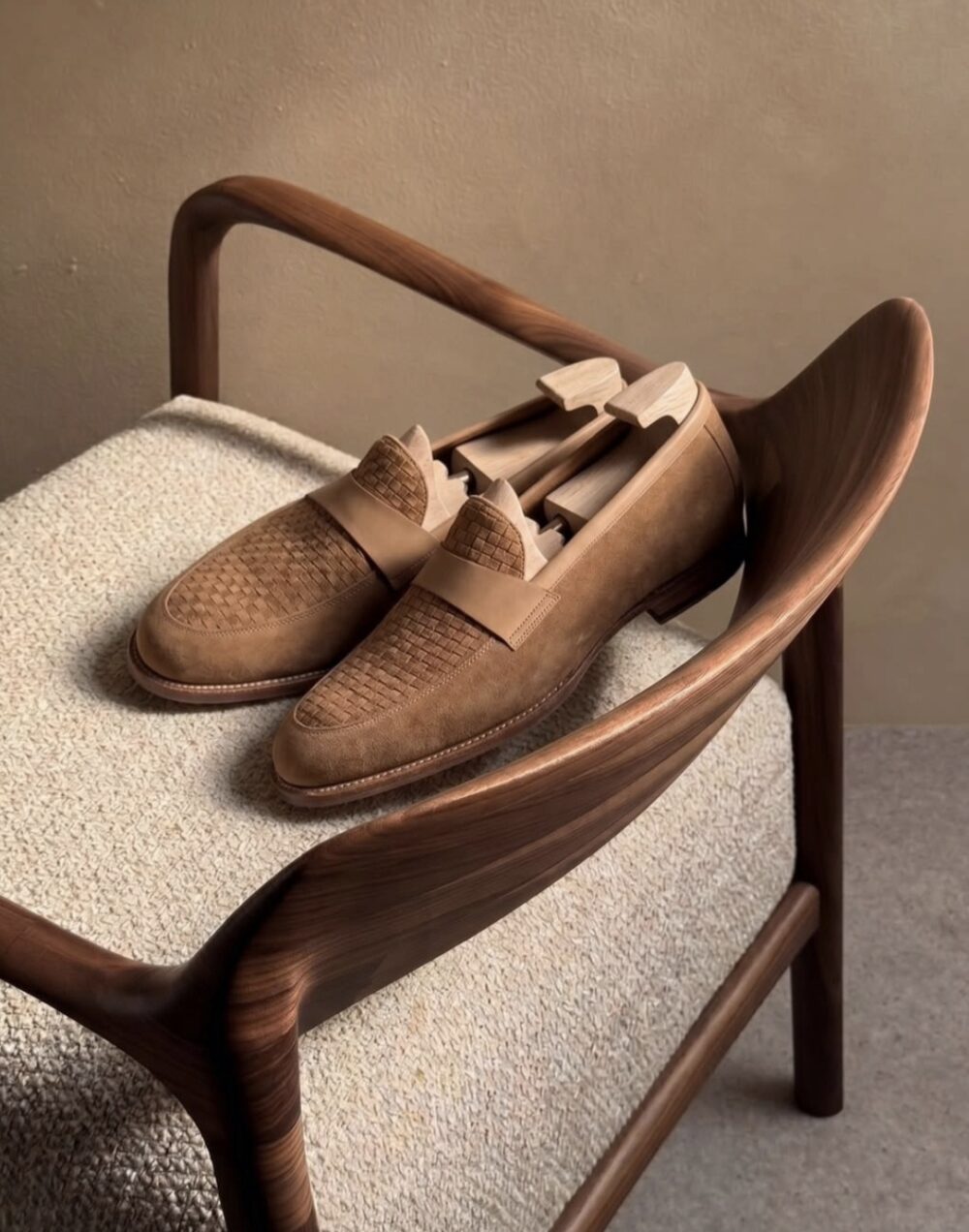A pair of brown suede Amalfi loafers with woven detailing rests on a wooden chair.