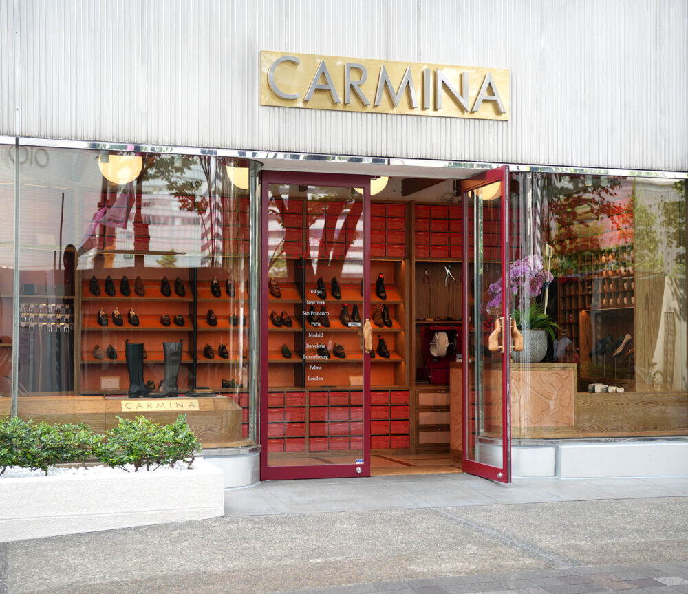 The image shows the entrance of a Carmina shoe store with a gold sign and red display boxes showcasing various shoes.