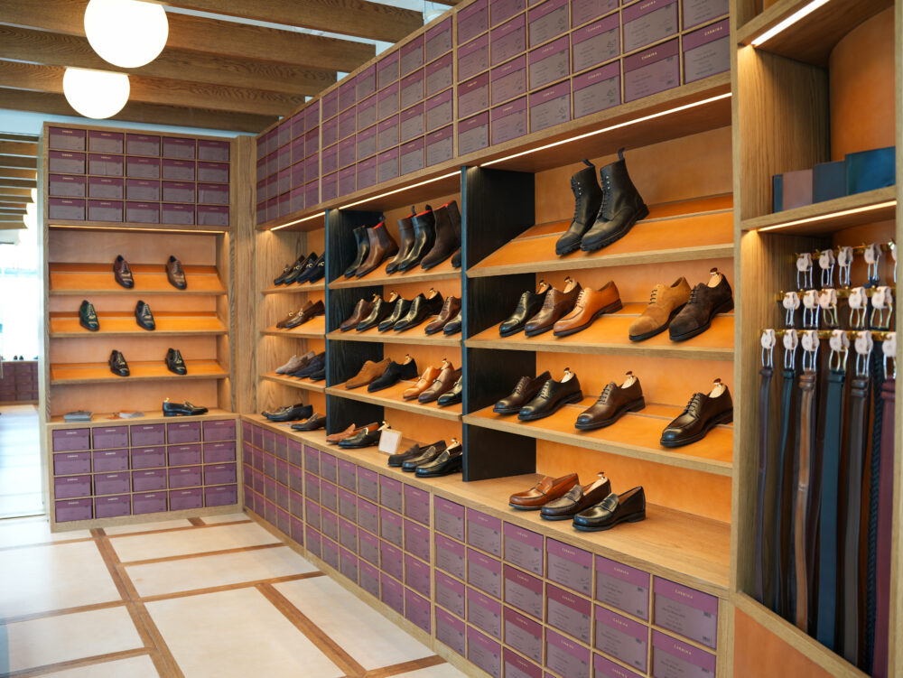 A display of various styles of shoes, including boots and dress shoes, arranged on wooden shelves with purple boxes below.