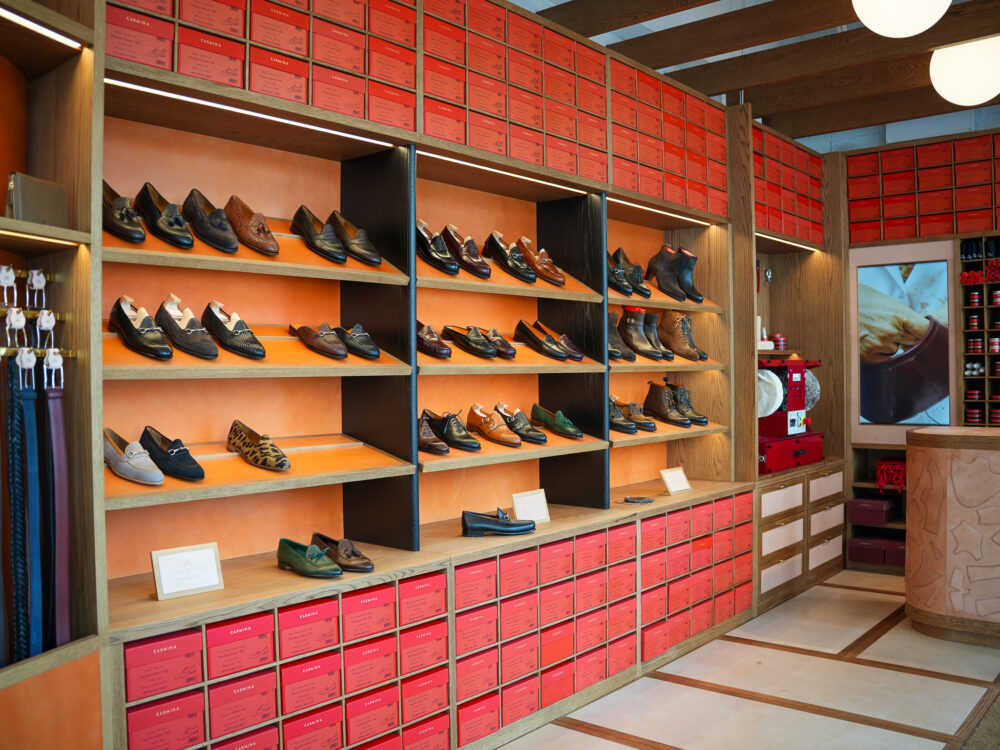 A display of various styles of Carmina shoes in shades of brown, black, and green on wooden shelves, with red shoe boxes below.