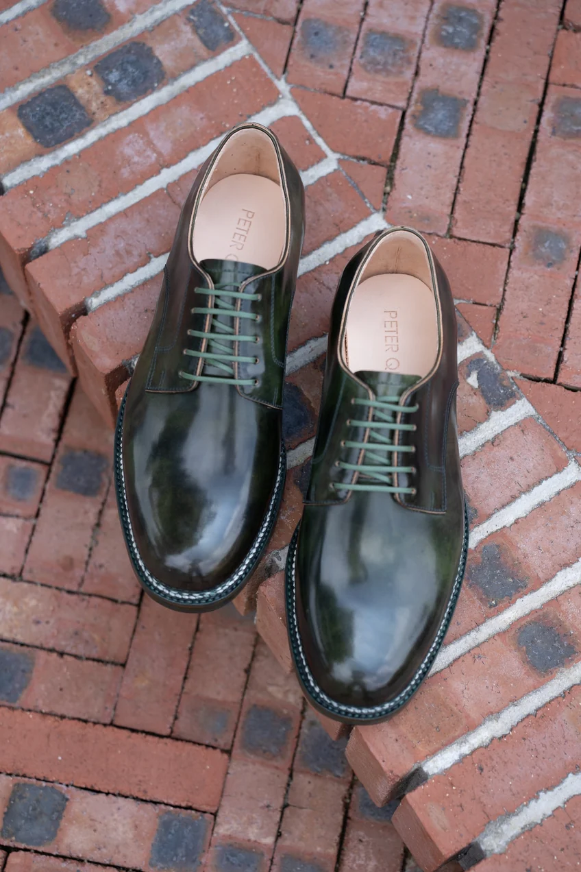 A pair of polished dark green cordovan shoes with brown laces, positioned on a brick surface.