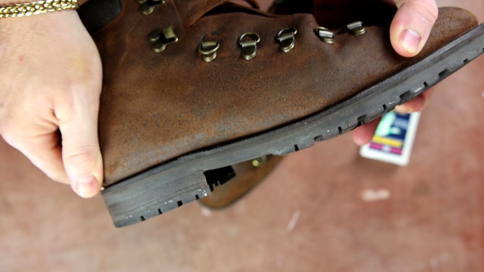 A person holds a worn brown leather boot, emphasizing its scuffed surface and showing Saphir Oiled Leather Cream in the background.