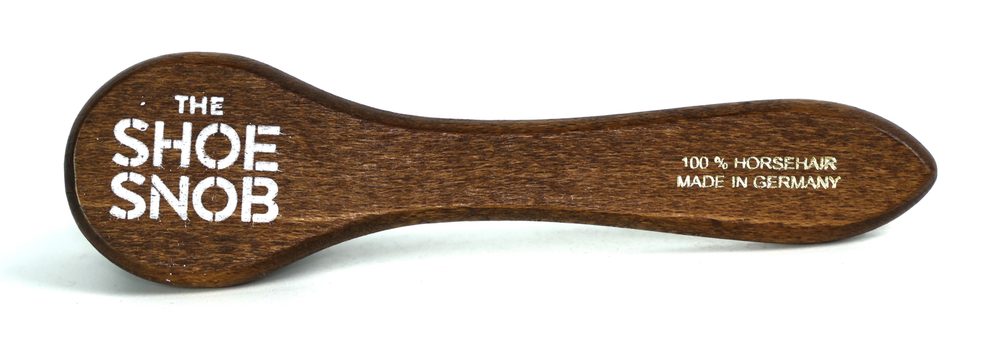 A wooden shoehorn labeled "The Shoe Snob," made from 100% horsehair and crafted in Germany.