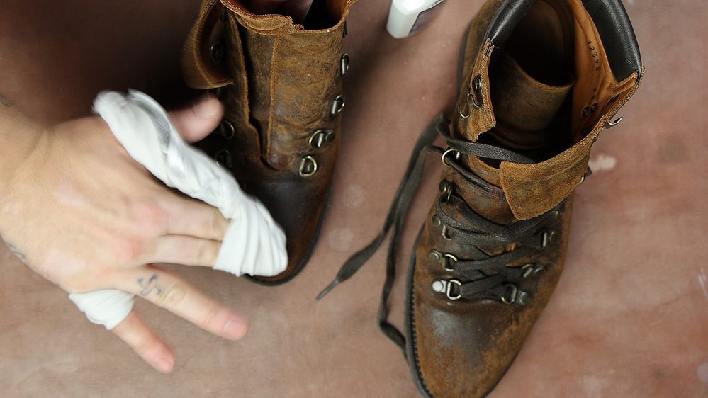 A hand is applying Saphir Oiled Leather Cream to a worn brown leather boot.
