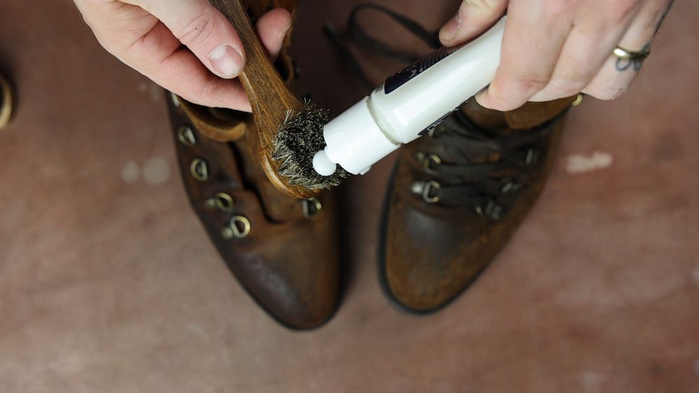 A person applies Saphir Oiled Leather Cream to a brush, preparing to restore brown leather boots.