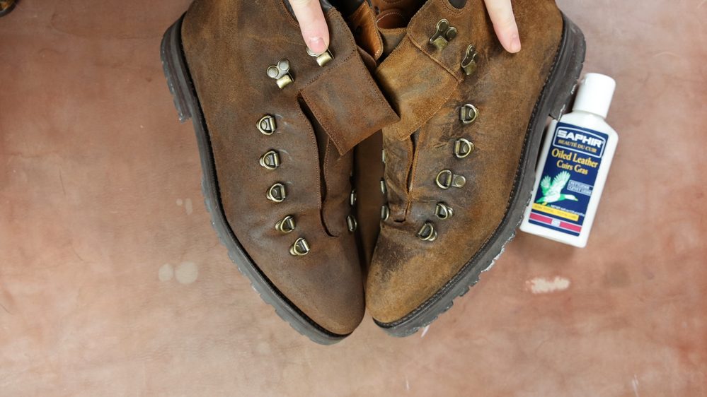A pair of worn brown leather boots with metal eyelets and Saphir Oiled Leather Cream positioned beside them.