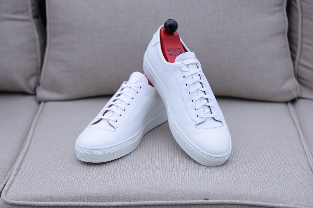 A pair of white leather Olympia White Calf Sneakers with red insoles and brown laces, positioned together.