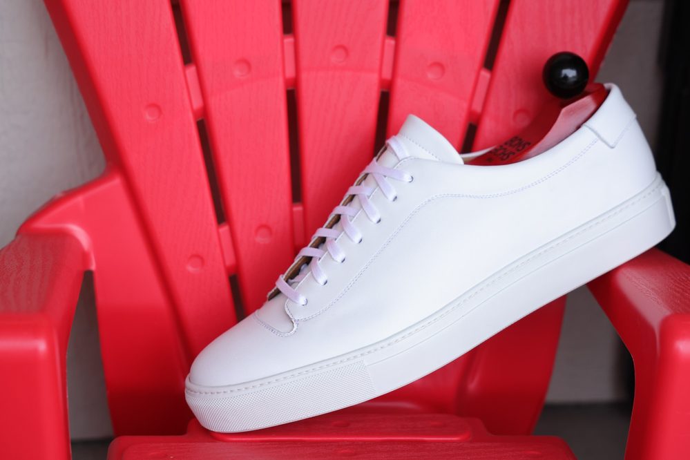 The image features a single white leather Olympia White Calf Sneaker resting on a bright red chair.