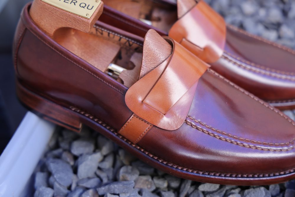A close-up of polished brown leather loafers with a tan leather strap and wooden shoe trees inside.