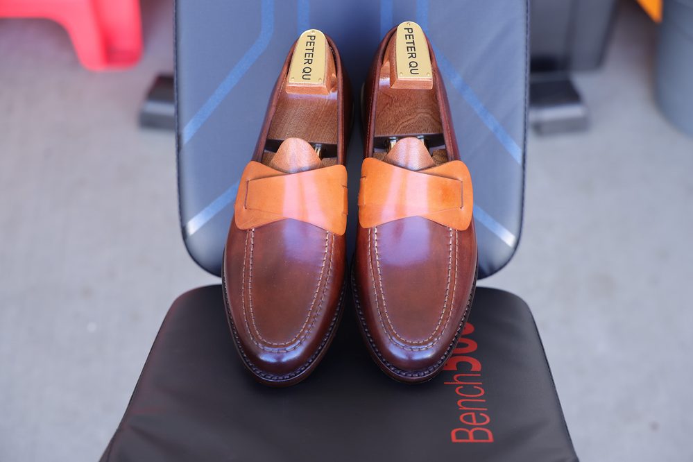 A pair of brown leather loafers with an orange leather bow detail on top, labeled "PETER QU."
