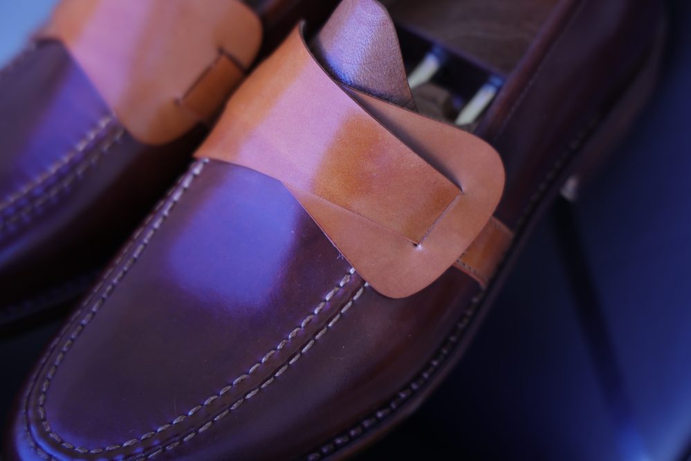 A close-up of a brown leather shoe featuring a tan leather strap across the front.