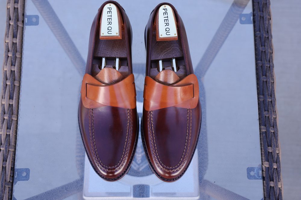 A pair of dark brown leather loafers with a light brown strap, featuring the brand name "Peter Qu" on the insoles.