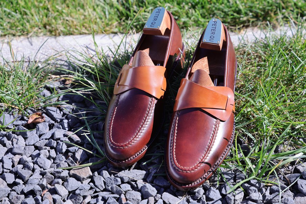 A pair of brown leather loafers with a tan strap across the front, featuring wooden shoe trees with "PETER QU" labels.