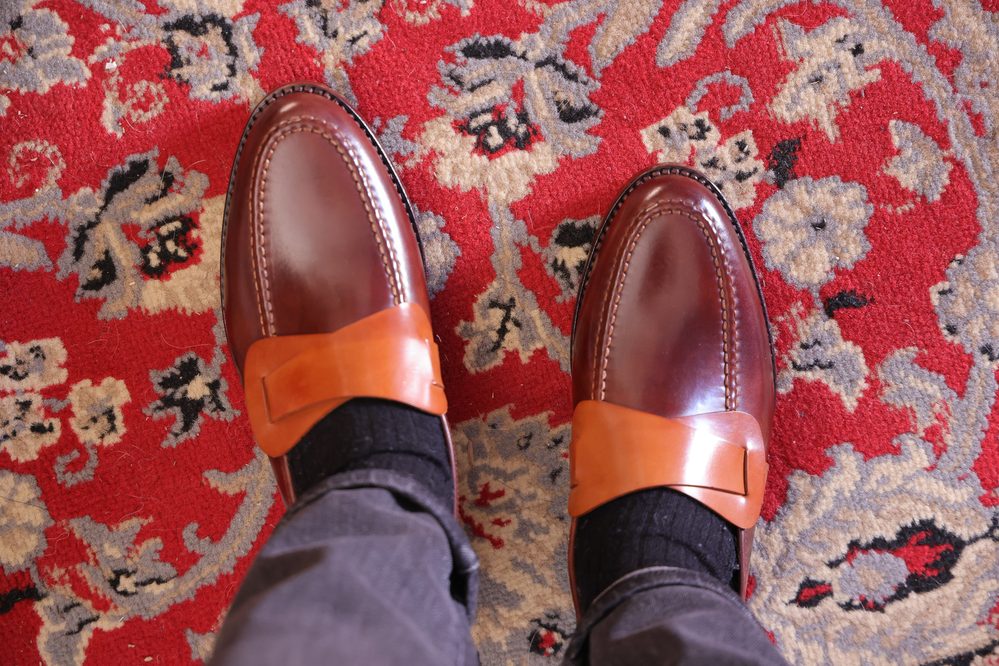 A close-up of two brown cordovan shoes with tan leather straps, worn with black socks.