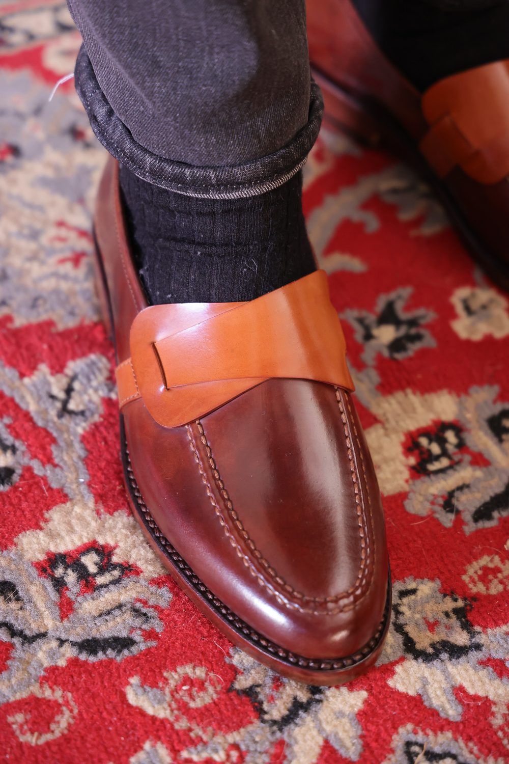 A close-up of a brown cordovan shoe with a tan leather strap, worn with black socks and dark jeans.