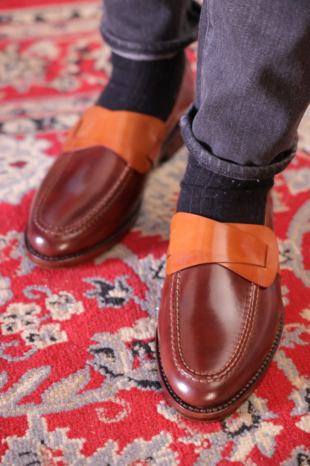 Brown cordovan loafers with a tan leather strap, worn with dark socks and denim jeans.