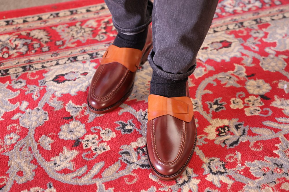 A close-up of brown leather shoes with an orange bow detail, worn with dark socks and jeans.