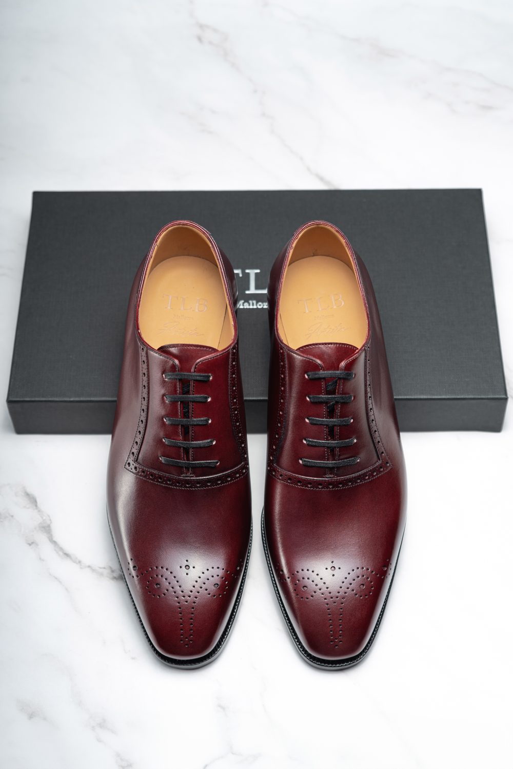 Burgundy leather dress shoes with decorative perforations, displayed on a black box.