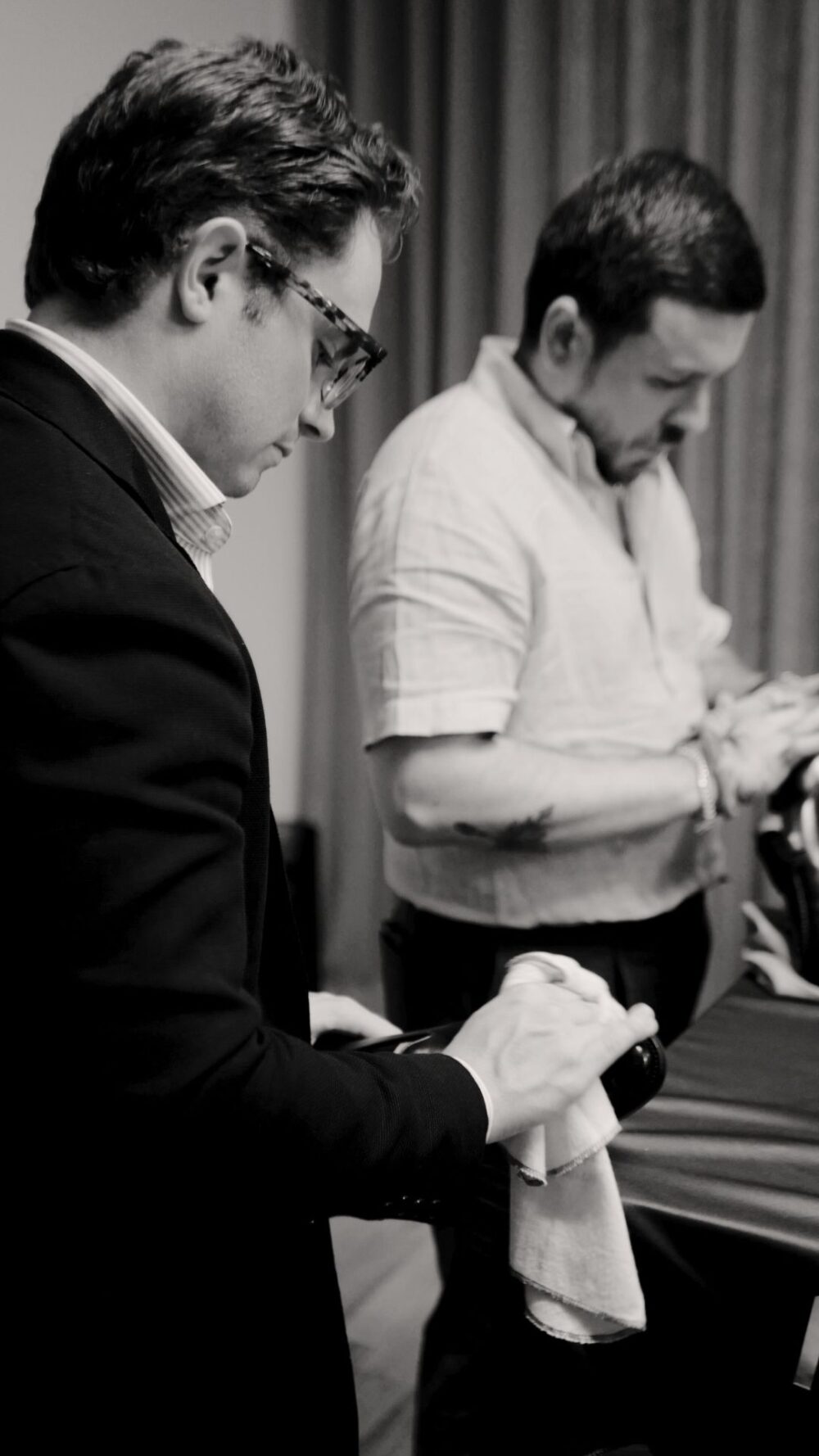 Two men are working on hats, one polishing with a cloth while the other examines a hat.