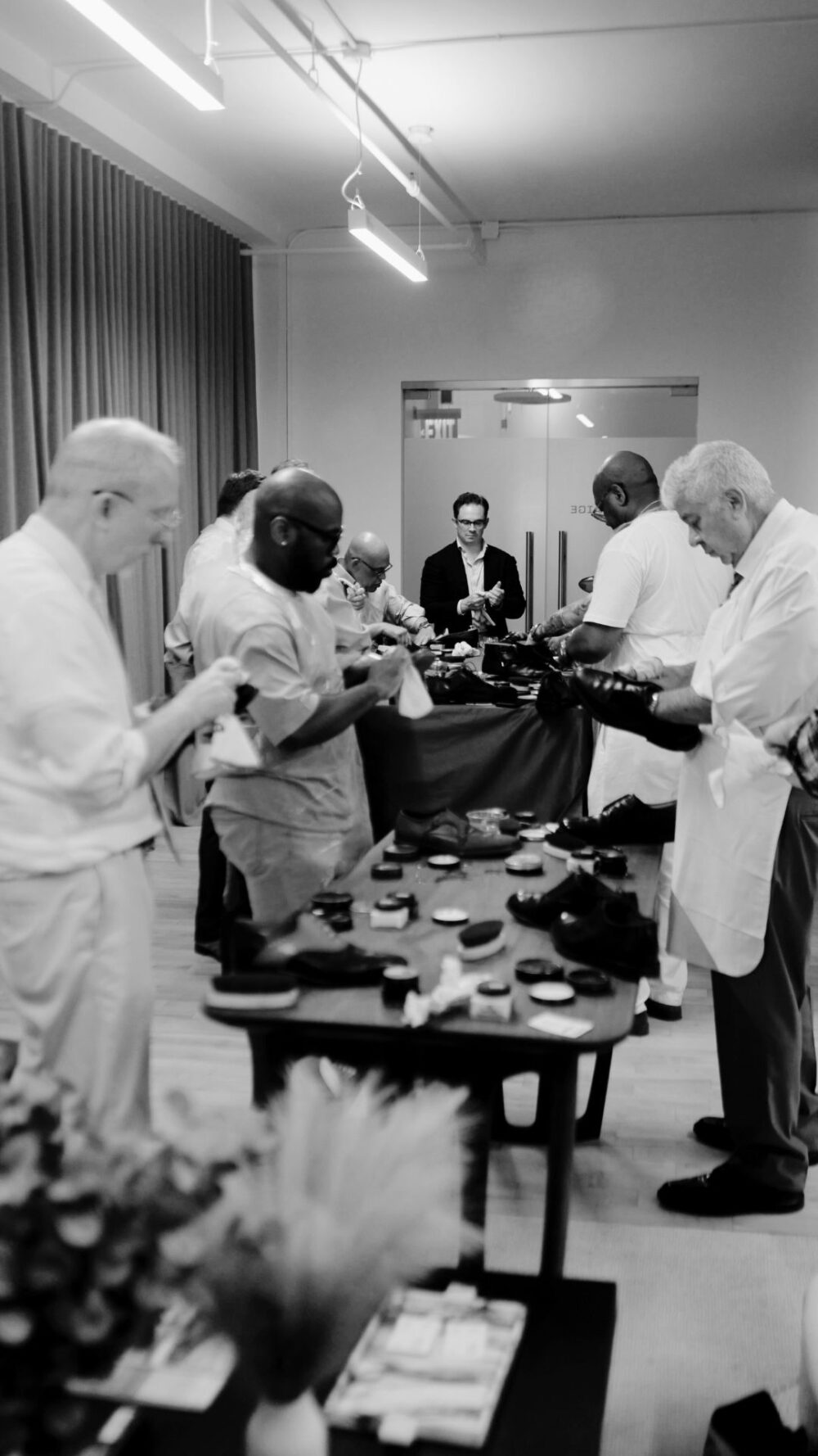 A group of people in white aprons examine hats and materials on a table during the Optimo Hat Factory Tour.