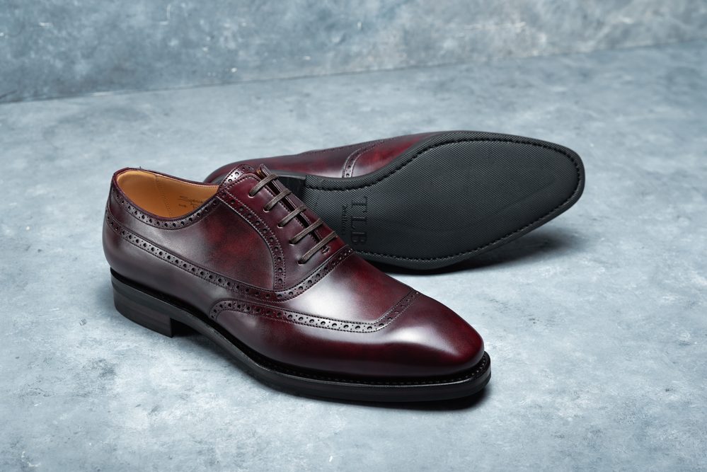 A pair of burgundy leather dress shoes with brogue detailing and a black rubber sole.