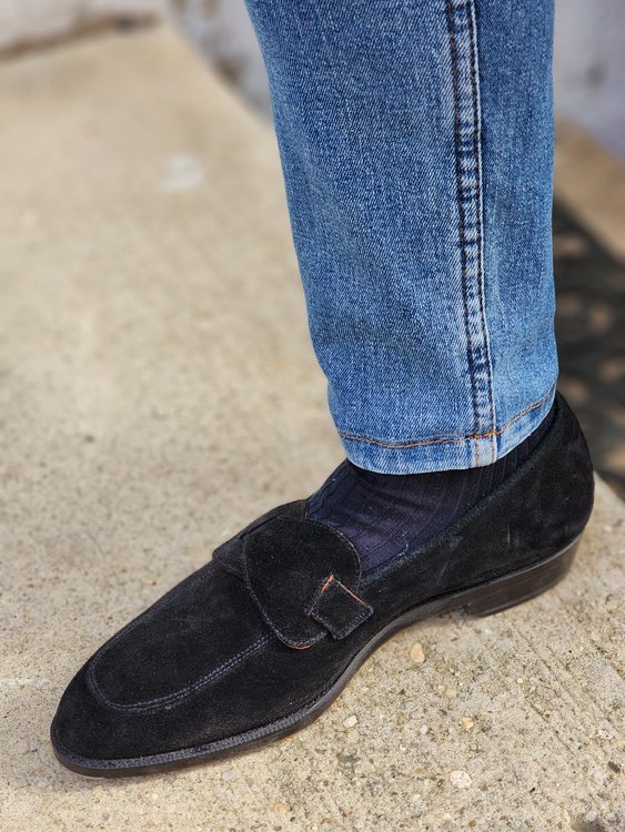 A close-up of a black suede shoe with a buckle, worn with blue denim jeans and dark socks.