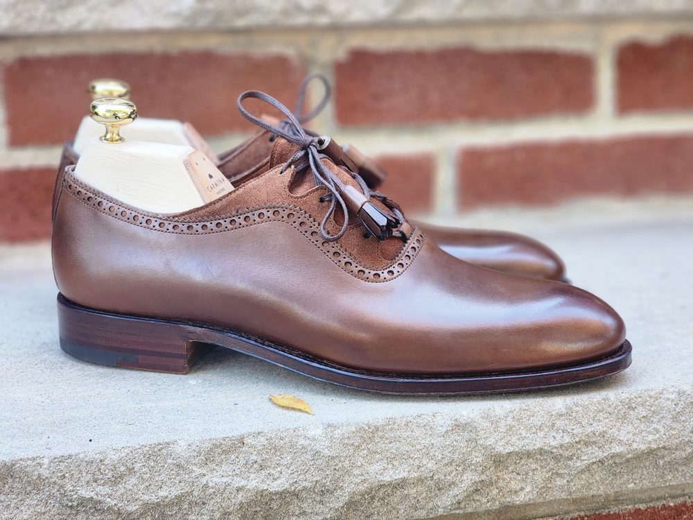 A pair of brown leather shoes with decorative perforations and wooden shoe trees, featured in the Carmina Bespoke Trunk Show.
