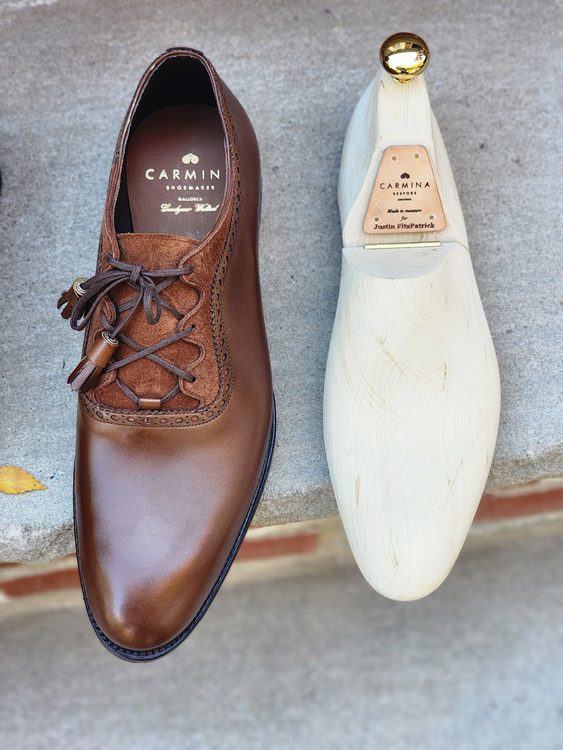 A polished brown leather shoe with intricate detailing is displayed next to a wooden shoe tree, featured in the Carmina Bespoke Trunk Show.