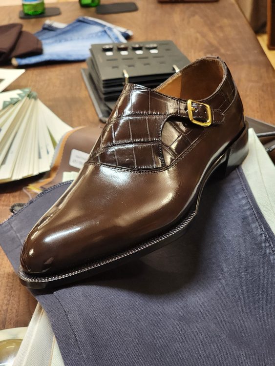A polished brown leather shoe with a crocodile nubuck detail and a gold buckle.