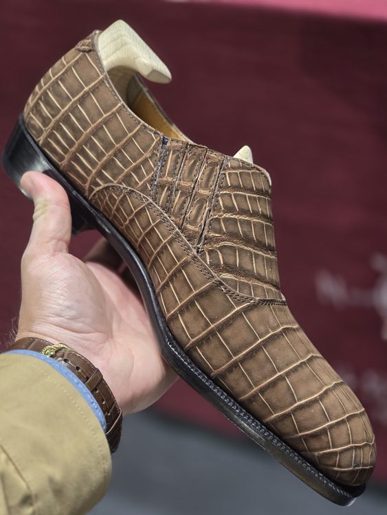 A brown crocodile nubuck shoe is held in a hand, showcasing its textured surface and sleek design.