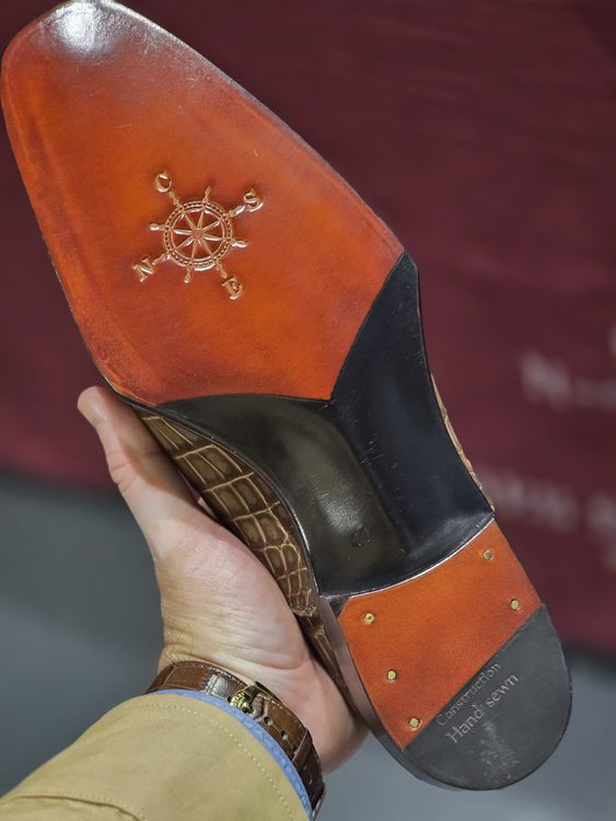 A hand holds the bottom of a shoe featuring a crocodile nubuck texture, with a tan leather sole embossed with a compass design.